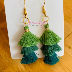 May include: A pair of green and teal tassel earrings with gold hooks. The earrings are layered with three tassels each.