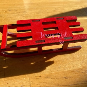 May include: A small, red wooden sled with dark brown accents. The sled has a classic design with runners, a seat, and a front bar. The text "Jersey Girl" is visible on the sled. The sled is a decorative item.