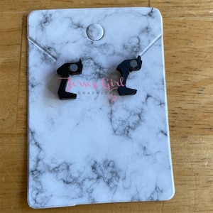 Puede incluir: Un par de pendientes de madera negra con forma de mano haciendo el dedo medio. Los pendientes están sobre un fondo de mármol blanco con el texto "Jersey Girl Graphics" impreso debajo.