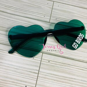 May include: Heart-shaped sunglasses with green lenses and black frames. The right lens has the text "GO BIRDS" in white. The sunglasses are on a light wood-grain surface. The text "Jersey Girl Graphics" is also visible.