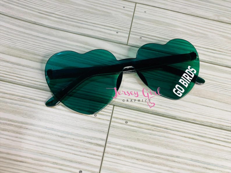 May include: Heart-shaped sunglasses with green lenses and black frames. The right lens has the text "GO BIRDS" in white. The sunglasses are on a light wood-grain surface. The text "Jersey Girl Graphics" is also visible.