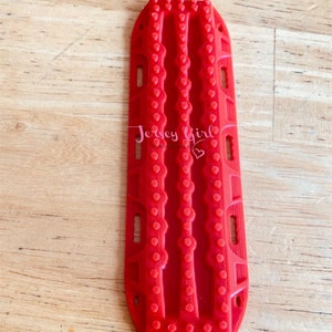 May include: Red plastic traction device with a textured surface and multiple rows of raised dots. The device is designed to help vehicles get unstuck in mud or snow.