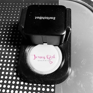 May include: A black SwitchBot device with a white sticker that says "Jersey Girl Graphics" with a pink heart.