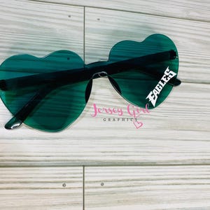 May include: Heart-shaped sunglasses with green lenses and black frames. The right lens features the word "EAGLES" in white. The sunglasses are set against a light wood-grain background. The text "Jersey Girl Graphics" is also visible.