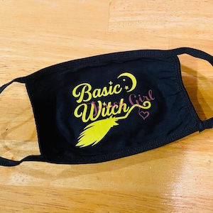 May include: Black cotton face mask with the text "Basic Witch Girl" in yellow lettering, a yellow broom, a crescent moon, and a pink heart.
