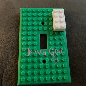 May include: Green light switch cover with a Lego brick design. The cover has the text "Jersey Girl Graphics" and a pink heart.
