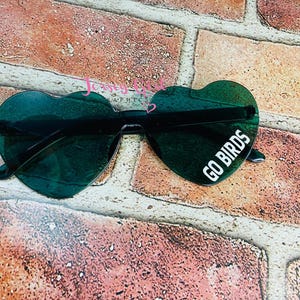 May include: Heart-shaped sunglasses with dark green lenses and black frames. The right lens features the white text "GO BIRDS". The sunglasses rest on a brick surface.