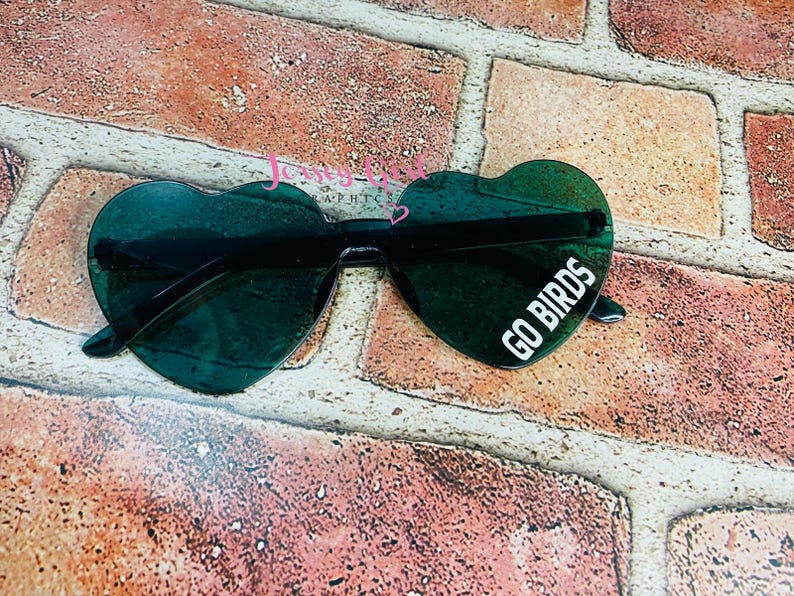 May include: Heart-shaped sunglasses with dark green lenses and black frames. The right lens features the white text "GO BIRDS". The sunglasses rest on a brick surface.