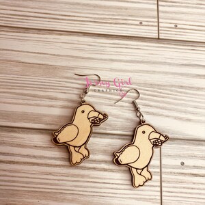 May include: A pair of wooden earrings featuring a cartoon seagull design. Each earring is painted in a light brown colour and has a silver hook.