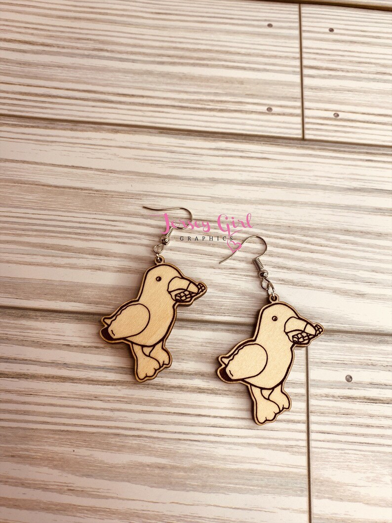 May include: A pair of wooden earrings featuring a cartoon seagull design. Each earring is painted in a light brown colour and has a silver hook.