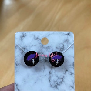 K&ouml;nnte beinhalten: Ein Paar Holz-Ohrstecker mit schwarzem Hintergrund und einem lila Blitz-Motiv. Die Ohrringe sind mit "Jersey Girl Graphics" beschriftet.