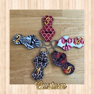 May include: Five different lip balm holders with keychains. Each holder has a different pattern: zebra print, leopard print, red and white geometric pattern, red lips print, and red and black splatter print. The holders are numbered 1 through 5.