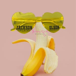 Peut inclure: Une banane pelée avec des lunettes de soleil jaunes en forme de cœur portant les inscriptions "JACKSON" et "OLSON" sur les verres. Les lunettes de soleil sont placées au-dessus de la banane sur un fond rose. L'image comprend également le texte "Jersey Girl Graphics".