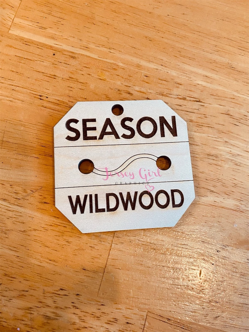 May include: Wooden sign with the text "SEASON" and "WILDWOOD" in black lettering. The sign has two holes for hanging and a pink graphic of a wavy line with the text "Jersey Girl Graphics" in pink.