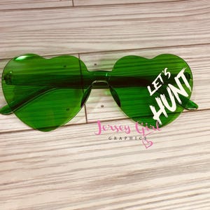 May include: Green heart-shaped sunglasses with the text "LET'S HUNT" in white. The sunglasses are on a light wood grain surface. The brand name "Jersey Girl Graphics" is also visible.