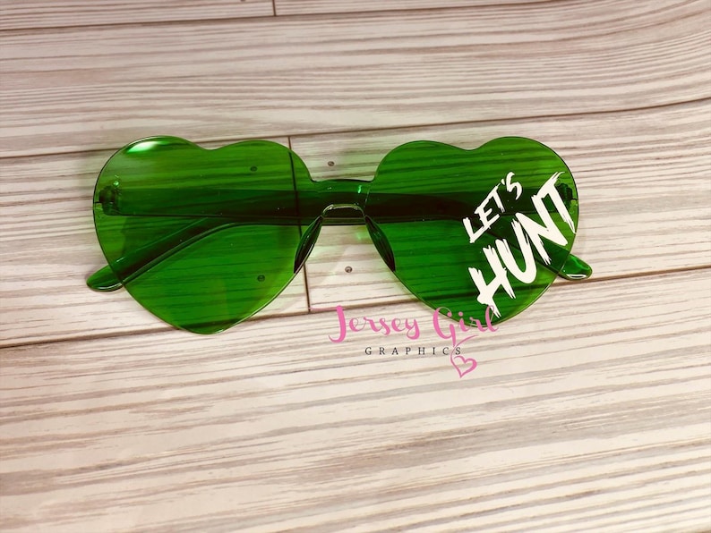 May include: Green heart-shaped sunglasses with the text "LET'S HUNT" in white. The sunglasses are on a light wood grain surface. The brand name "Jersey Girl Graphics" is also visible.
