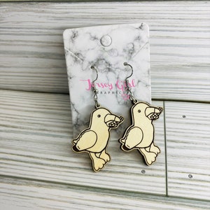 May include: A pair of wooden earrings featuring a cartoon bird design. The birds are light brown and have a small amount of detail in black. The earrings are hanging from silver hooks.