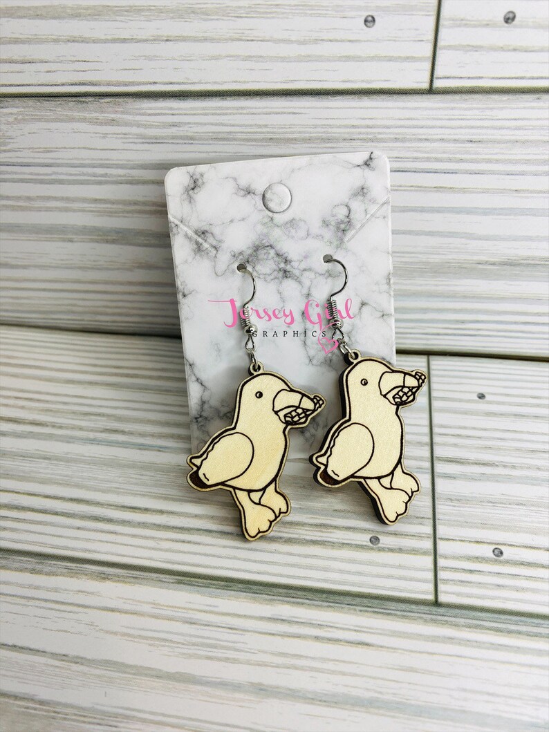 May include: A pair of wooden earrings featuring a cartoon bird design. The birds are light brown and have a small amount of detail in black. The earrings are hanging from silver hooks.