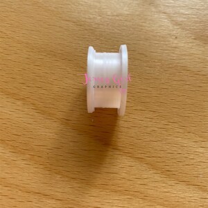 May include: A small, white plastic spool with a central opening, resting on a light brown wooden surface. The spool has a cylindrical shape with raised edges. The text "Jersey Girl Graphics" is visible in pink.