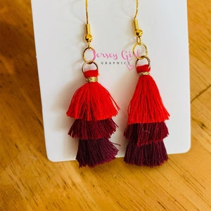 May include: A pair of red and burgundy tassel earrings with gold hooks. The tassels are layered and have a fringe design.