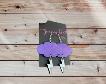 May include: Purple glitter cloud earrings with silver lightning bolt charms. The earrings are hanging from a black card with the text "Jersey Girl Graphics" on it.