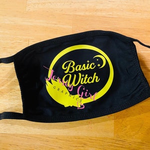 May include: Black face mask with a yellow circle and the text "Basic Witch" in a yellow script font. The text "Jersey Girl Graphics" is also in yellow script font.
