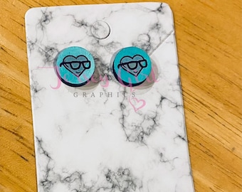 May include: A pair of teal blue heart-shaped earrings with black outlines of a heart wearing glasses. The earrings are on a white marble background.