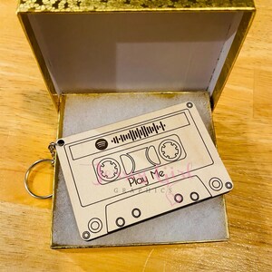 May include: A wooden keychain shaped like a cassette tape with the text "Play Me" and a Spotify logo. The keychain is in a gold gift box.