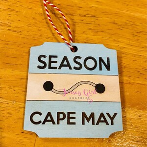 May include: A blue and white wooden ornament with the text "SEASON CAPE MAY" and a small gray line with two black dots. The ornament has a red and white string attached to it.