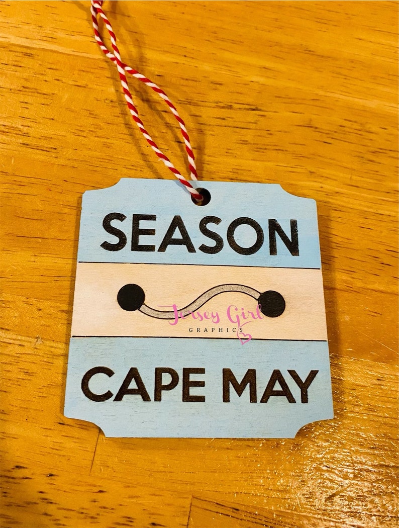 May include: A blue and white wooden ornament with the text "SEASON CAPE MAY" and a small gray line with two black dots. The ornament has a red and white string attached to it.