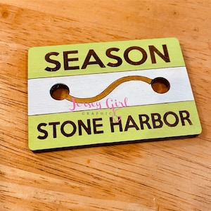May include: A green and white wooden sign with the text "SEASON STONE HARBOR" and a gold wavy line. The sign has two holes in it.