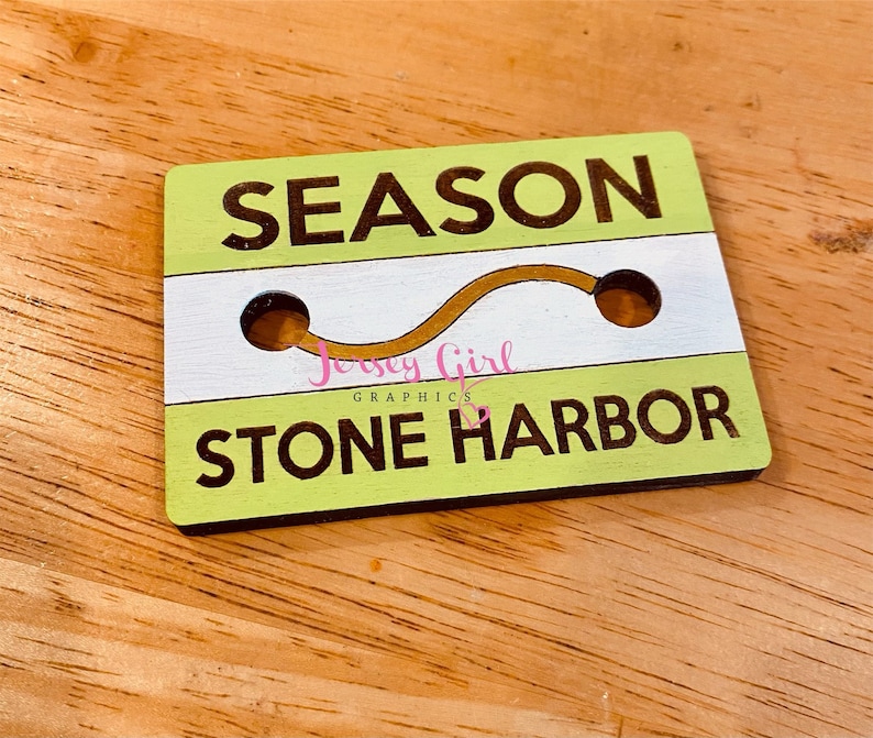 May include: A green and white wooden sign with the text "SEASON STONE HARBOR" and a gold wavy line. The sign has two holes in it.