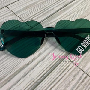 May include: Heart-shaped sunglasses in a dark green hue. The lenses feature the text "GO BIRDS" in white. The sunglasses are set against a light wood grain background.