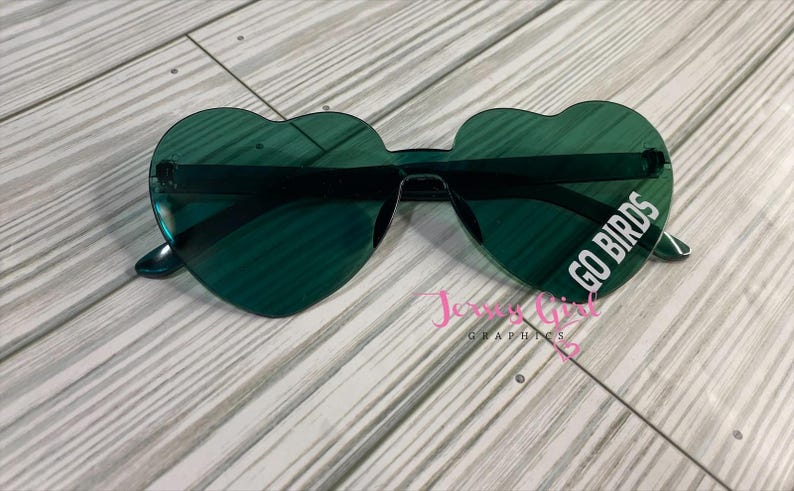May include: Heart-shaped sunglasses in a dark green hue. The lenses feature the text "GO BIRDS" in white. The sunglasses are set against a light wood grain background.
