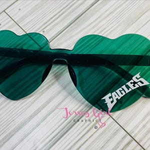 May include: Heart-shaped sunglasses in a vibrant green hue. The lenses feature the word "EAGLES" in white, with a black frame. The sunglasses are set against a light wood-grain background.