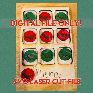 May include: A digital file for a laser cut dinosaur game. The game board is made of wood and has nine squares. Each square has a red or green circle with a dinosaur inside. The text "DIGITAL FILE ONLY!" is printed on the board. The text "Nora" is printed below the game board. The text ".SVG LASER CUT FILE" is printed below the name.