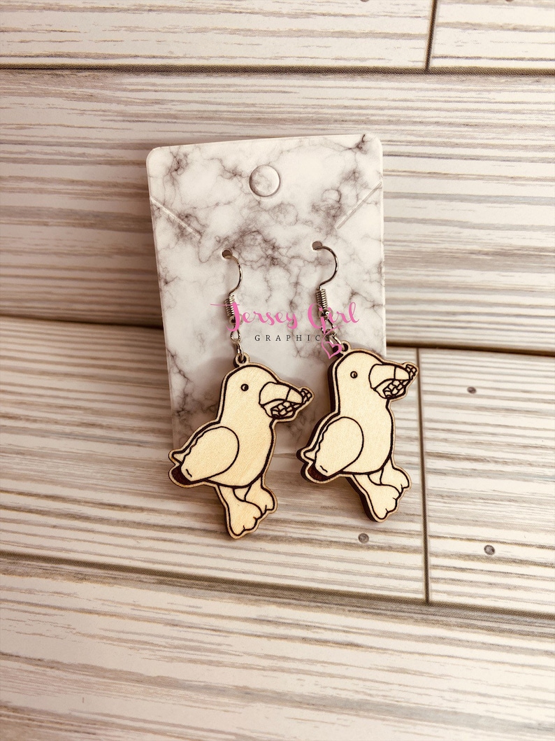 May include: A pair of wooden earrings featuring a cartoon bird design. The earrings are light brown with black outlines and are hanging from silver hooks.