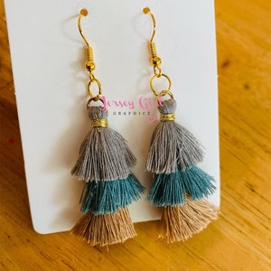 May include: A pair of gold tassel earrings with three layers of tassels in gray, teal, and beige.