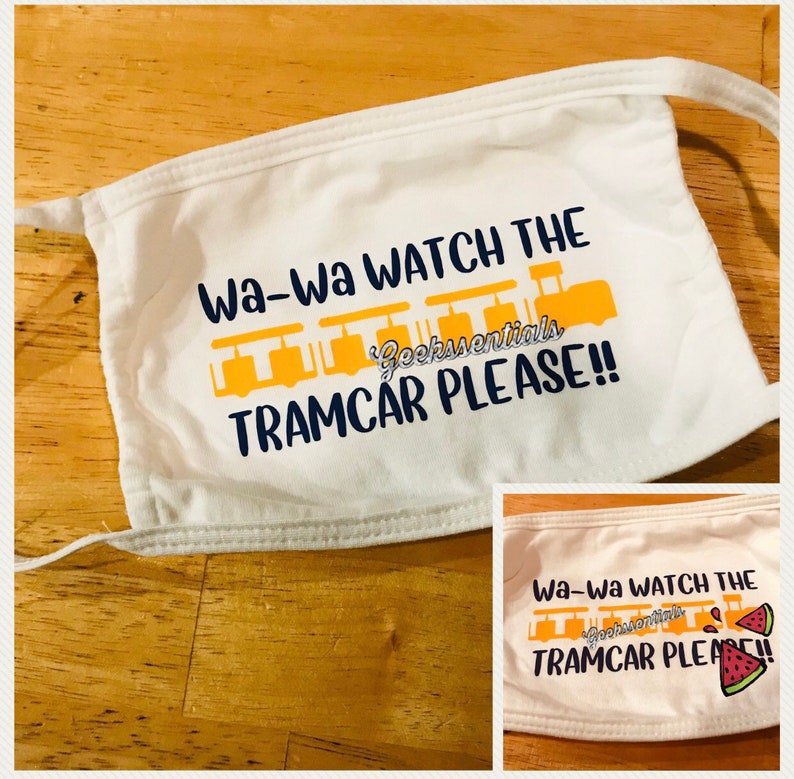 Puede incluir: M&aacute;scara facial blanca con el texto "Wa-Wa WATCH THE TRAMCAR PLEASE!!" en letras amarillas y azules. El texto est&aacute; acompa&ntilde;ado por un gr&aacute;fico de un tranv&iacute;a amarillo.