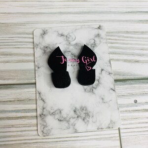 May include: Two black felt earrings in the shape of a pointed heart. The earrings are displayed on a white marble background with the text "Jersey Girl Graphics" in pink.
