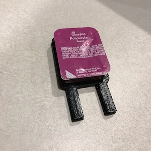 May include: A single-serving packet of Chick-fil-A Polynesian Sauce. The packet is pink and white and has the Chick-fil-A logo and the words "Polynesian Sauce" printed on it. The packet is sitting on a black plastic holder.