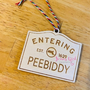 May include: A wooden Christmas bauble with the text "Entering Peebiddy Est. 1629 Jersey Girl Graphics" and a small outline of the state of New Jersey.