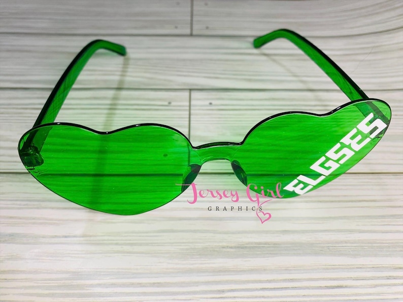 May include: Green heart-shaped sunglasses with the word "ELGSES" in white block letters on the right lens. The sunglasses have a clear frame and arms. The image also includes the text "Jersey Girl GRAPHICS".