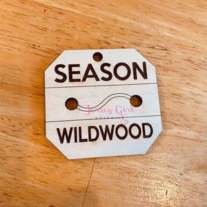 May include: Wooden sign with the text "SEASON" and "WILDWOOD" in black lettering. The sign has two holes for hanging and a pink graphic of a wavy line with the text "Jersey Girl Graphics" in pink.