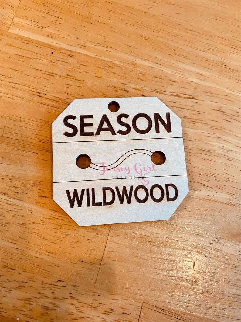 May include: Wooden sign with the text "SEASON" and "WILDWOOD" in black lettering. The sign has two holes for hanging and a pink graphic of a wavy line with the text "Jersey Girl Graphics" in pink.