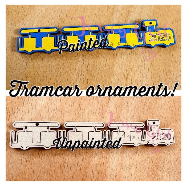 May include: Two wooden tramcar ornaments, one painted blue and yellow with the year 2020 on the last car, and the other unpainted. The painted ornament has the word "Painted" on it, and the unpainted ornament has the word "Unpainted" on it. Both ornaments have a hole at the top for hanging.