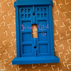 May include: Blue plastic light switch cover shaped like the TARDIS from Doctor Who. The cover has a white light switch in the center and the text "Jersey Girl Graphics" in pink.