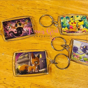 May include: Four clear plastic keychains with different Pokemon characters. Each keychain features a colorful illustration of a Pokemon character against a background. The keychains are attached to silver key rings.