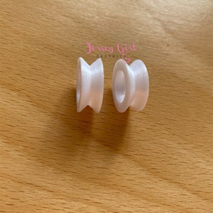 May include: Two white, spool-shaped objects with a central hole and a flared, ridged design. The objects are placed on a light brown wooden surface. The text "Jersey Girl Graphics" is visible in the upper right corner.
