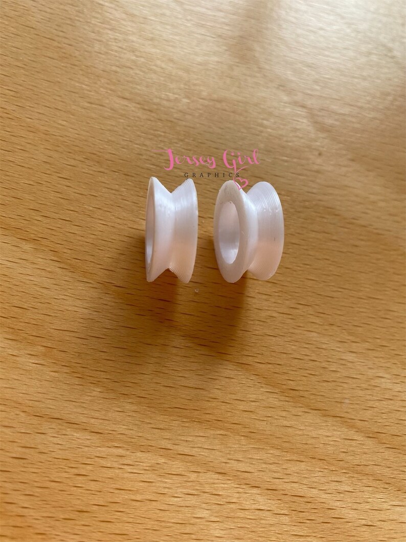 May include: Two white, spool-shaped objects with a central hole and a flared, ridged design. The objects are placed on a light brown wooden surface. The text "Jersey Girl Graphics" is visible in the upper right corner.